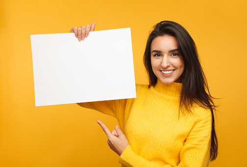 Young content model in yellow sweater holding and pointing at blank paper in hands smiling at camera.