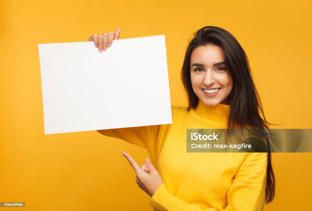 Charming brunette pointing at blank paper Young content model in yellow sweater holding and pointing at blank paper in hands smiling at camera. Holding Stock Photo
