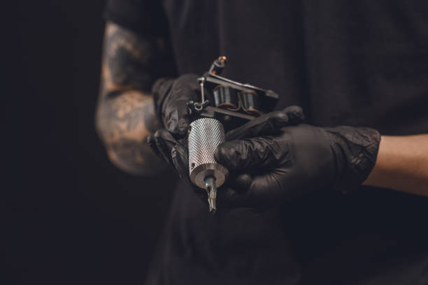 mani maschili in guanti che tengono macchina per tatuaggi isolata su nero - tatuare foto e immagini stock