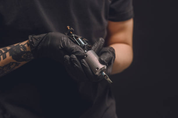istock Tattoo master in gloves holding ink machine isolated on black 936432992