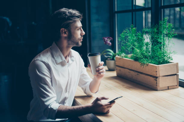 istock Time for inspiration and coffee. Young freelancer is thinking, he is dreaming and enjoying, sitting in a modern cafe near the window, holding his mobile phone in a hand 936443284