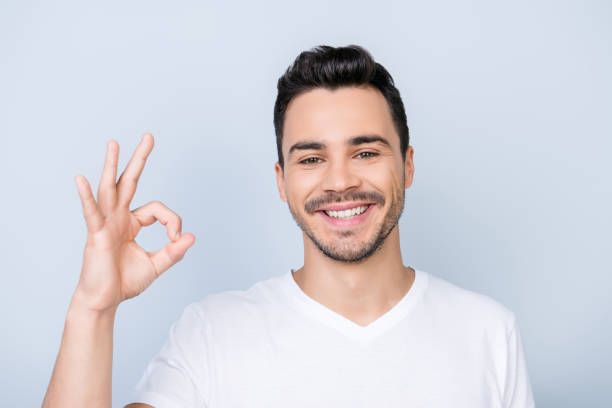 istock Young cheerful successful guy on the pure light blue background is smiling, wearing casual outfit and is showing ok sign 936443876