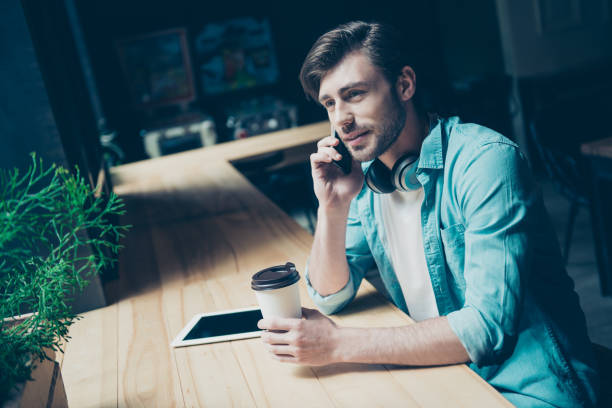 istock Young stylish hipster student is resting after studying, having hot tea and nice conversation on his phone, smiling, in jeans shirt 936444208