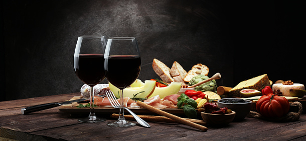 Italian antipasti wine snacks set. Cheese variety, Mediterranean olives, pickles, Prosciutto di Parma with melon, salami and wine in glasses over black grunge background