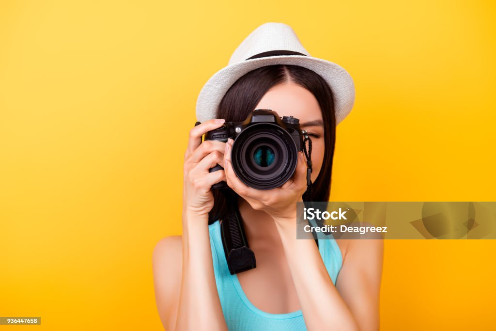 Feche-se de um fotógrafo fazendo um tiro em uma câmera digital durante as férias. Ela está vestindo roupa casual de verão e um chapéu, em fundo amarelo brilhante - Foto de stock de Fotógrafo royalty-free