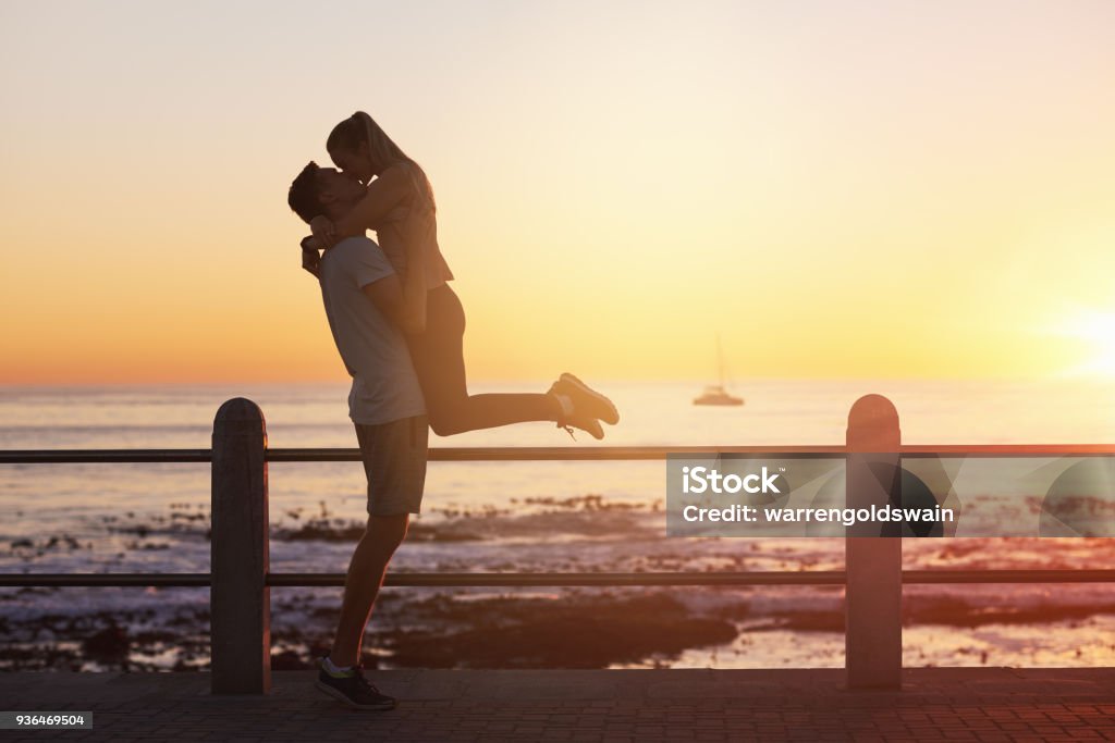 Young woman jumps into boyfriend's arms and gives him a kiss at sunset Young woman jumps on boyfriend and gives him a kiss at sunset, romantic passionate moment on date, golden sun flare Couple - Relationship Stock Photo