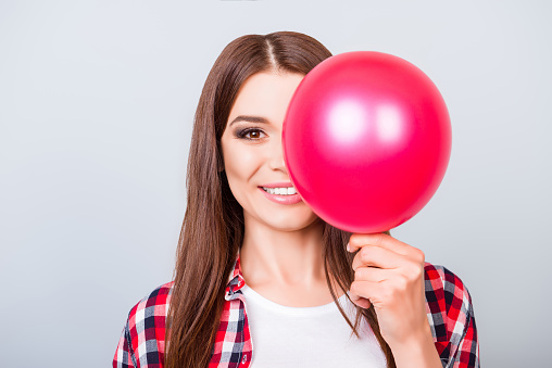 Cute brown haired young lady in casual wear is hiding half of her face with red balloon, standing on grey background, with beaming smile