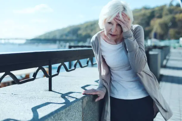 Need to take my pills. Exhausted older lady leaning on a barrier and touching her forehead while suffering from a terrible headache outdoors.