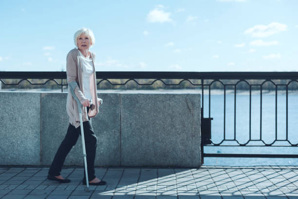 Lack of energy. Unhappy elderly woman looking into vacancy while leaning on crutches and walking along the river.