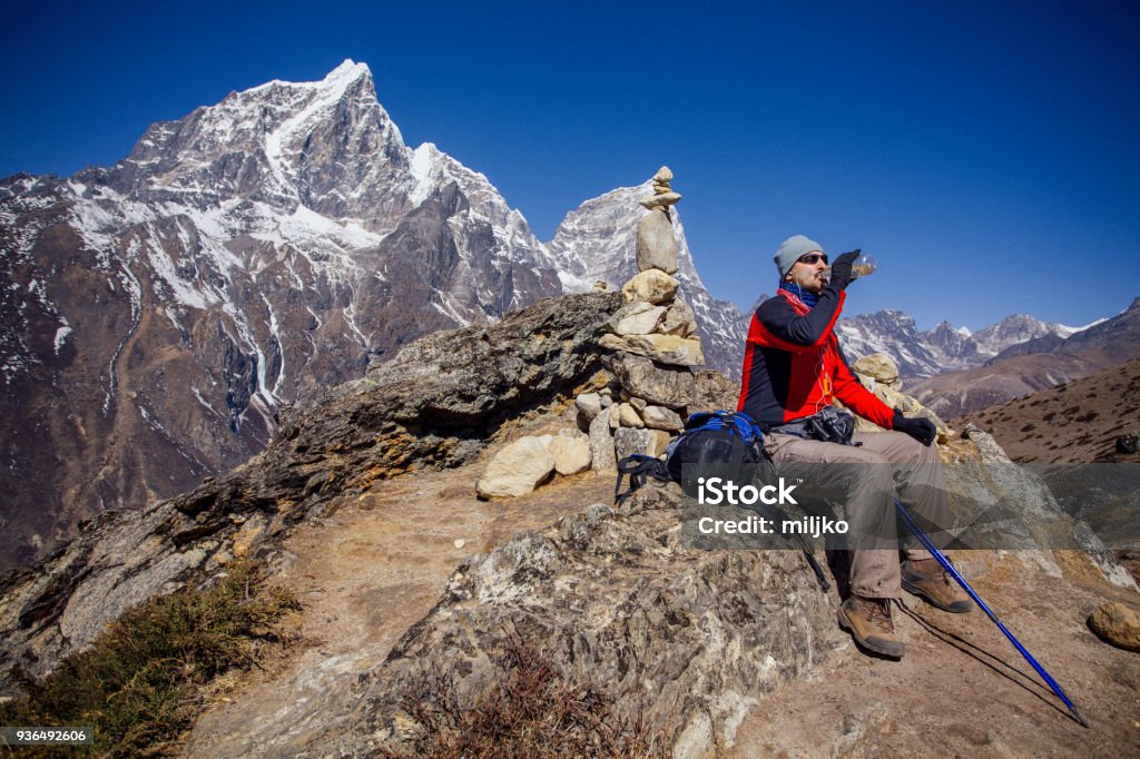 Hiking on Himalayas on Everest trek One man hiker and adventurer relaxing while sitting on the stone and drinking water from bottle. He is located on Himalayas, high above sea level, among some of the highest mountains on the world Achievement Stock Photo