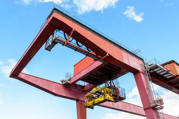 View from below of a red container gantry crane showing the spreader used for transloading intermodal containers from container barge in a river port, against blue sky.