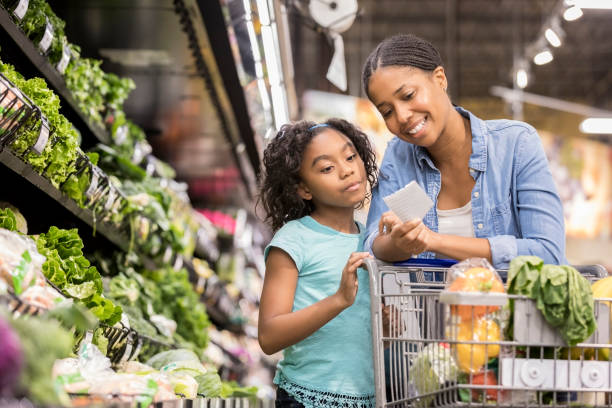 Mother and daughter grocery shop together using list A smiling mid adult woman stands in the produce section of her supermarket and reaches down to show her serious elementary age daughter the next items on a paper shopping list. supermarket stock pictures, royalty-free photos & images