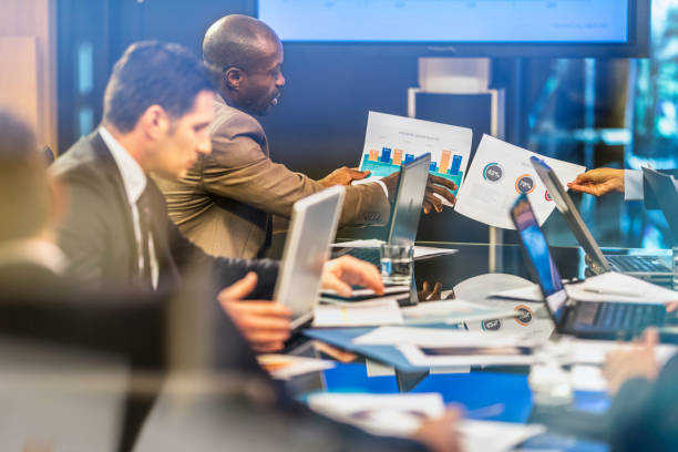 Comparing graphs on a board meeting African American businessman showing infographic to his invisible colleague also holding infographic, on a board meeting at conference table. hedge fund stock pictures, royalty-free photos & images