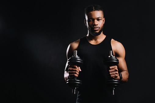 muscular young african american sportsman holding dumbbells and looking at camera isolated on black