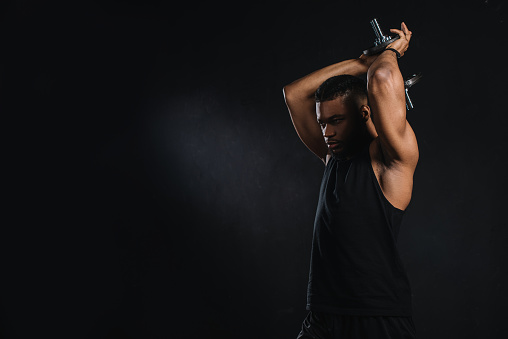 muscular young african american sportsman exercising with dumbbell isolated on black