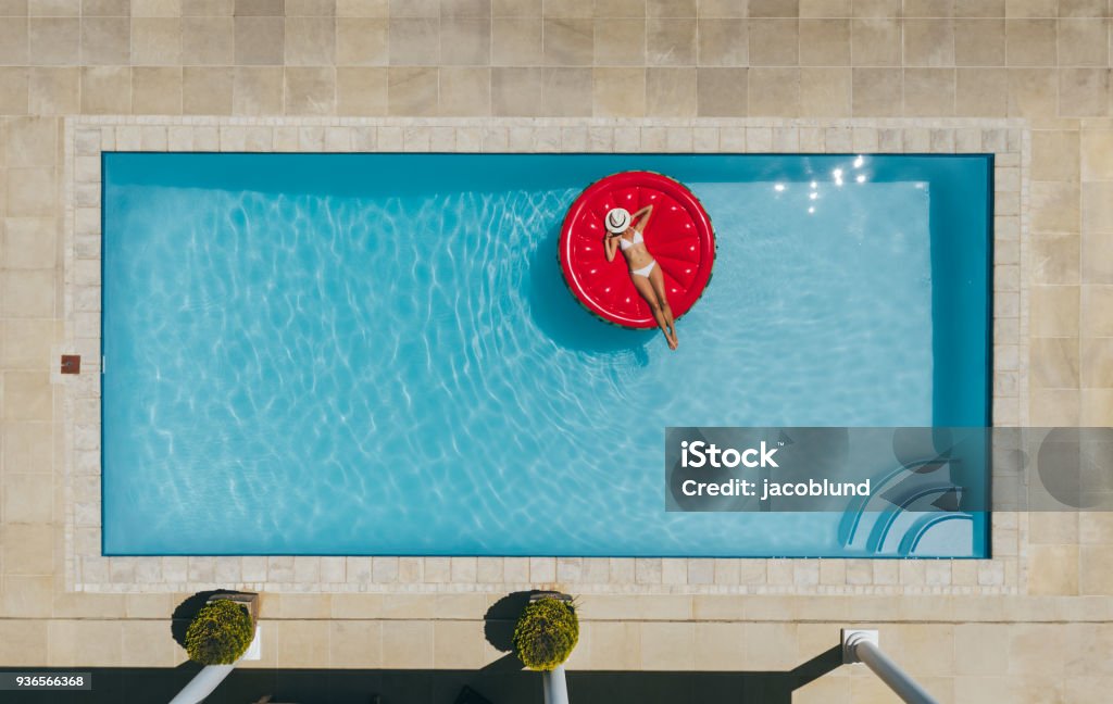 Woman sunbathing on inflatable mattress in pool. Aerial view of female in bikini lying on a floating mattress in swimming pool with her face covered with hat. Top view of woman sunbathing on inflatable mattress in pool. Swimming Pool Stock Photo