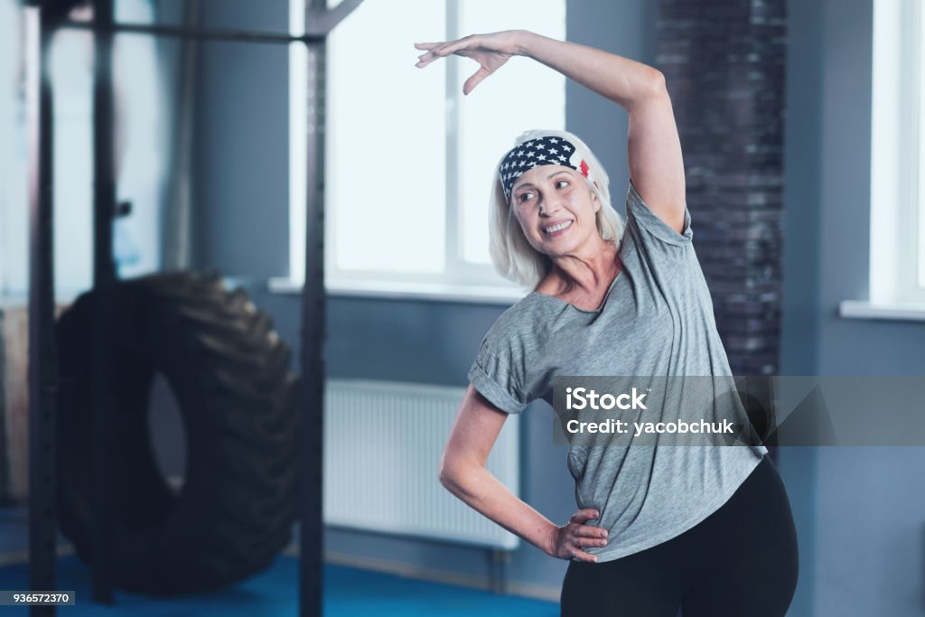 Cheerful mature lady stretching during training session Sport is life. Positive minded woman grinning broadly and looking somewhere while doing side bend exercises in a gym. Senior Adult Stock Photo