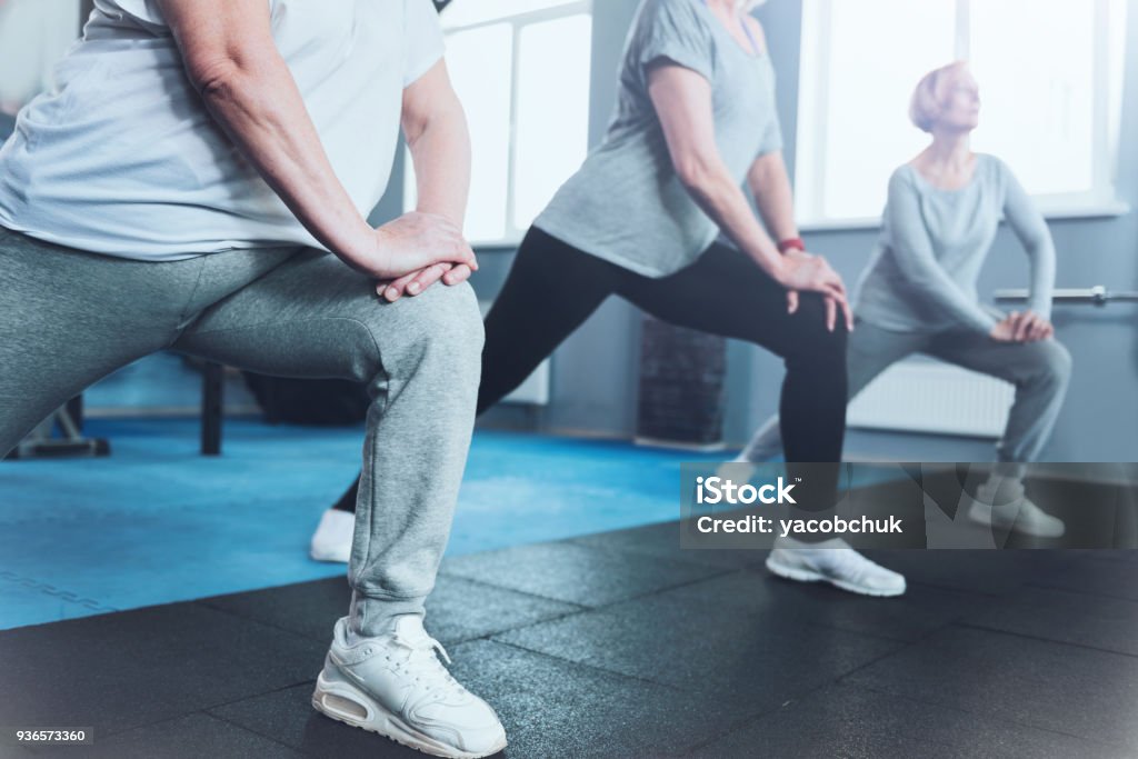 Close up look on retired women lunging at fitness club Forget about your joint pain. Scale up shot of a group of senior ladies standing in a row and doing lunging exercises while all three training in a gym. Senior Adult Stock Photo