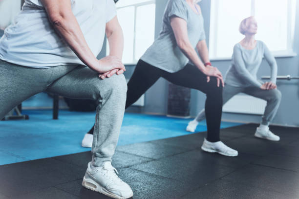 Close up look on retired women lunging at fitness club Forget about your joint pain. Scale up shot of a group of senior ladies standing in a row and doing lunging exercises while all three training in a gym. knee stock pictures, royalty-free photos & images