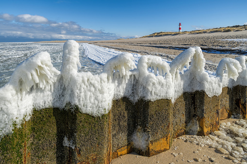 Icecapped iron groyne and lighthouse on sand dune in winter on island Sylt, Germany.