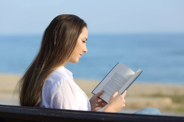 istock Woman reading a book on a bench on the beach 936584162