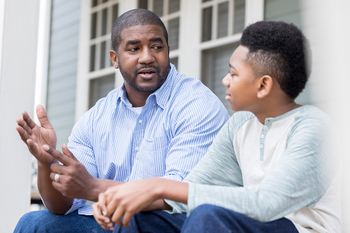 Caring mid African American father gestures as he gives advice to his preteen son. They are looking at one another. They are sitting on the front porch of their home.