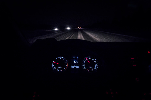 Car driver point of view perspective looking through dark night windshield above digital display electronic dashboard speedometer, odometer, and other gauges at snowy, slippery expressway and blurry traffic vehicles up ahead.