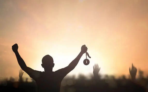 Silhouette winner hand holding gold medal reward against blurred sport stadium sunset background