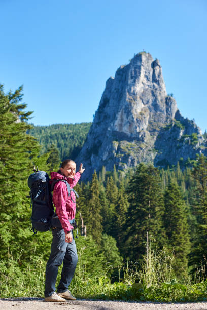 istock Hiker girl in modern outfit with blue backpack pointing with a hand on big stone cliff. Young hiker woman in pink jacket and gray pants enjoying view of high mountain in sunny warm weather. 936689628
