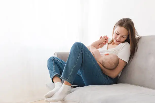 Mother is breastfeeding her kid sitting against light window background. Mom is suckling baby boy at home