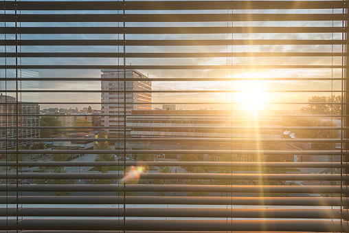 The blinds in a hotel room in amsterdam have turned down. The slats are tilted. The morning sun shines through them. It's blinding. The sun's rays penetrate the room. Beautiful morning atmosphere in an unknown city.