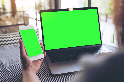 Mockup image of a woman holding blank mobile phone while using laptop with blank green screen on table