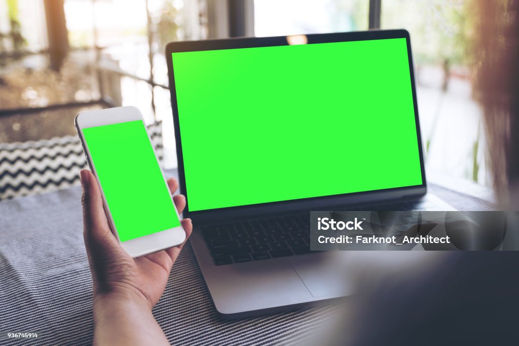 Mockup image of a woman holding blank mobile phone while using laptop with blank green screen on table Laptop Stock Photo