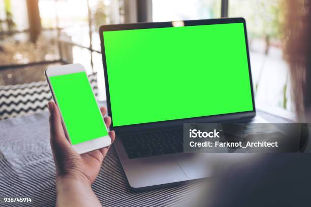 Mockup Image Of A Woman Holding Blank Mobile Phone While Using Laptop With Blank Green Screen On Table Stock Photo - Download Image Now