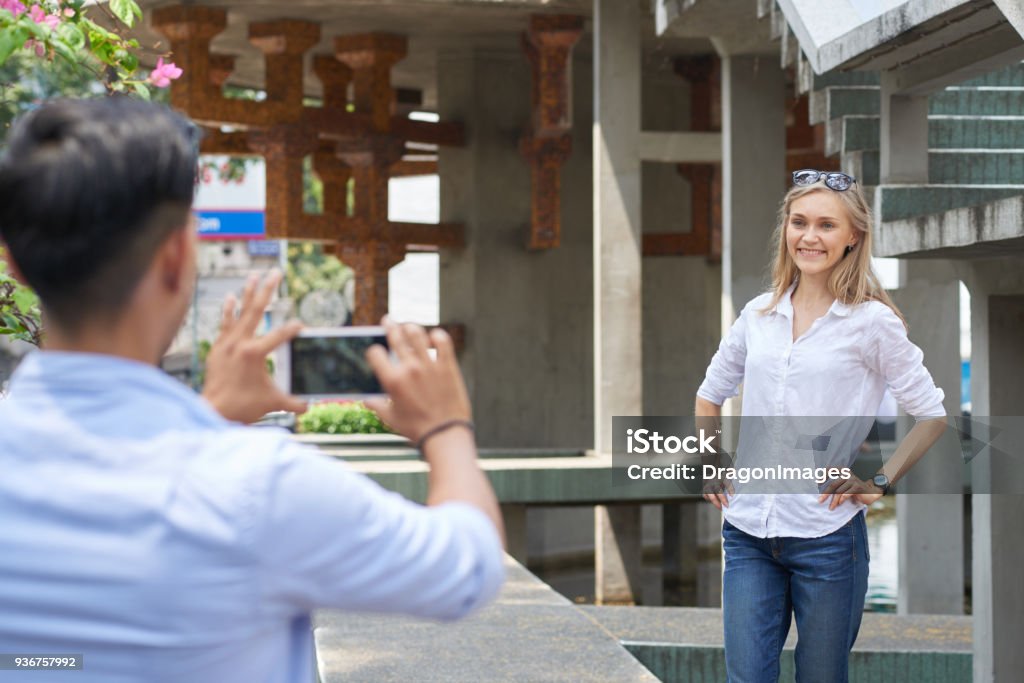 Posing for boyfreind Asian man photographing his beautiful Caucasian girlfriend with smartphone Adult Stock Photo
