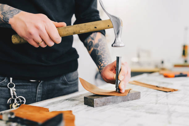 istock Close up of a leather craftsman working with leather using hammer. Working process in the leather workshop. Man holding crafting tool and working. 936804424