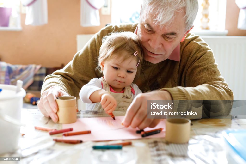 Cute little baby toddler girl and handsome senior grandfather painting with colorful pencils at home. Grandchild and man having fun together Cute little baby toddler girl and handsome senior grandfather painting with colorful pencils at home. Grandchild and man having fun together. Family and generation in love. Grandparent Stock Photo
