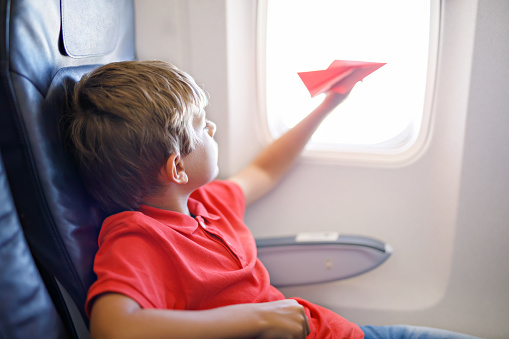 Little kid boy playing with red paper plane during flight on airplane. Child sitting inside aircraft by a window. Family going on vacation.