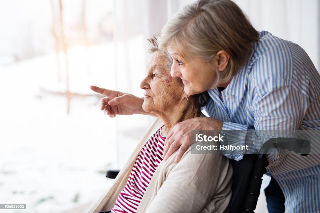 Senior women with wheelchair at home. An elderly mother with her daughter at home looking out of the window. A carer assisting a disabled senior woman in wheelchair. Senior Adult Stock Photo