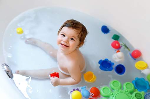 Cute adorable baby girl taking foamy bath in bathtub. Toddler playing with bath rubber toys. Beautiful child having fun with colorful gum toys and foam bubbles.