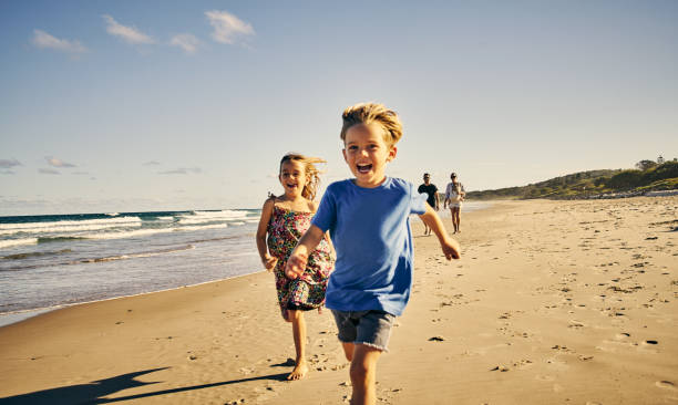Leading the way to a day of fun Shot of two adorable little children running at the beach with their parents in the background family trips and holidays stock pictures, royalty-free photos & images