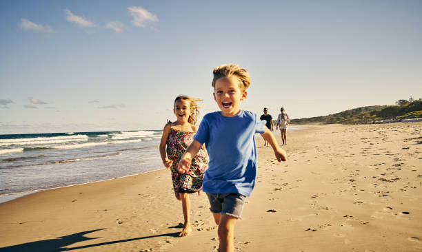 Leading the way to a day of fun Shot of two adorable little children running at the beach with their parents in the background beach fun stock pictures, royalty-free photos & images