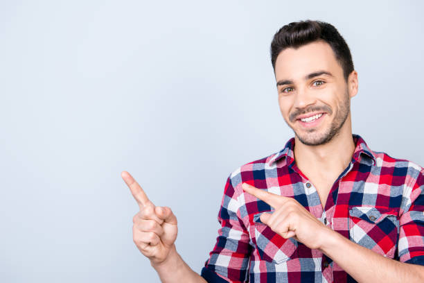 istock Young cheerful successful entrepreneur on the pure light blue background is smiling, wearing casual outfit and is pointing on a copyspace with his fingers 936840738