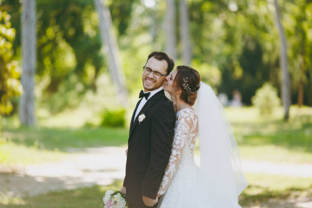 beau mariage photosession. la mariée à une dentelle blanche robe avec une longue plume, voile et bouquet les piqûres de l’oreille son fiancé dans un costume noir dans un grand jardin verdoyant sur une journée ensoleillée weathery - veil bride lace married photos et images de collection