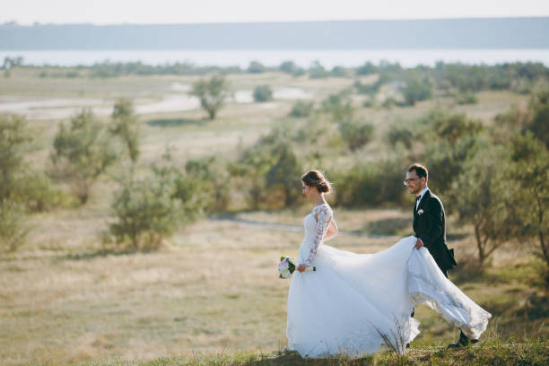 beau mariage photosession. beau marié dans un costume noir et une jeune mariée en robe de dentelle blanche avec coiffure exquise promenade autour du grand terrain vert dans le contexte d’arbres et d’arbustes - veil bride lace married photos et images de collection