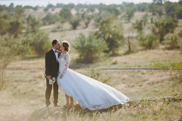 beau mariage photosession. beau marié dans un costume noir et une jeune mariée en robe de dentelle blanche avec coiffure exquise promenade autour du grand terrain vert dans le contexte d’arbres et d’arbustes - veil bride lace married photos et images de collection