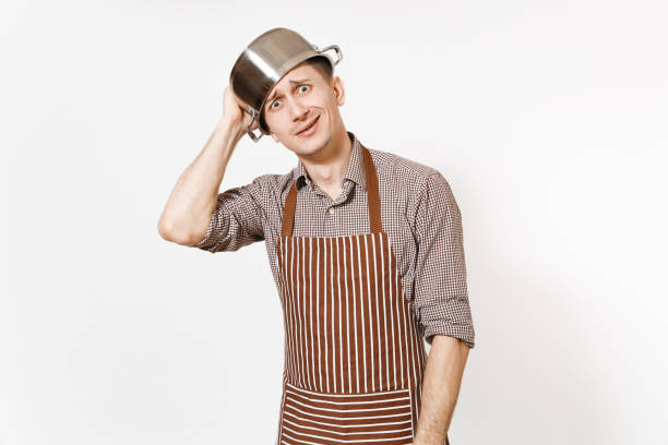 istock Fun man in striped apron with silver stainless glossy aluminium empty stewpan, pan or pot on head isolated on white background. Male housekeeper or houseworker. Kitchenware, dishes, cuisine concept. 936849840