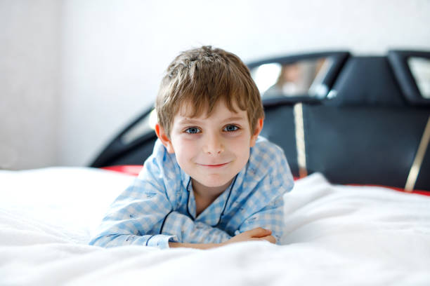 愛らしいハッピー子供男の子カラフルなナイトウェアで白いベッドで寝た後。小学生パジャマ パーティーを祝っていると、カメラを見て - portrait germany party bed ストックフォトと画像