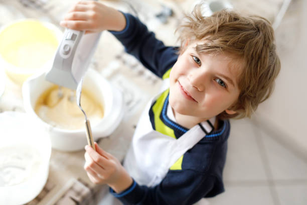 beautiful funny blond little kid boy baking chocolate cake and tasting dough in domestic kitchen - cake making mixing eggs imagens e fotografias de stock