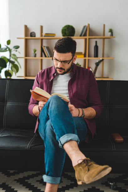 istock Young male in glasses sitting on leather couch and reading book 936885886